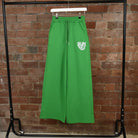 Green SUNBAE Sweat Pants with a logo on a hanger against a brick wall.