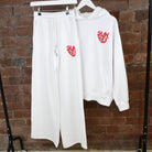 White hoodie and pants with red text on hangers against a brick wall.