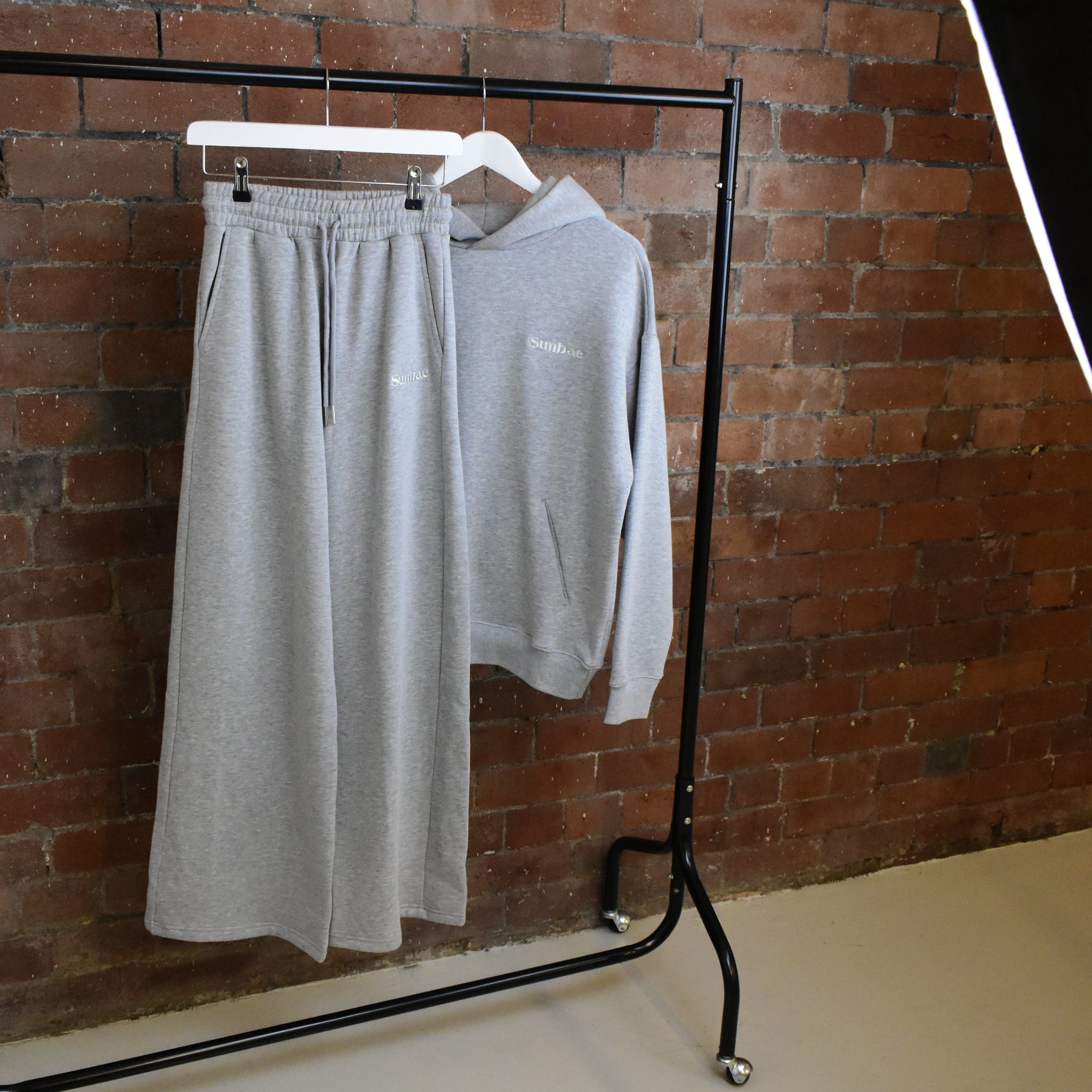 Grey SUNBAE sweatpants and hoodie on a black coat rack against a brick wall.
