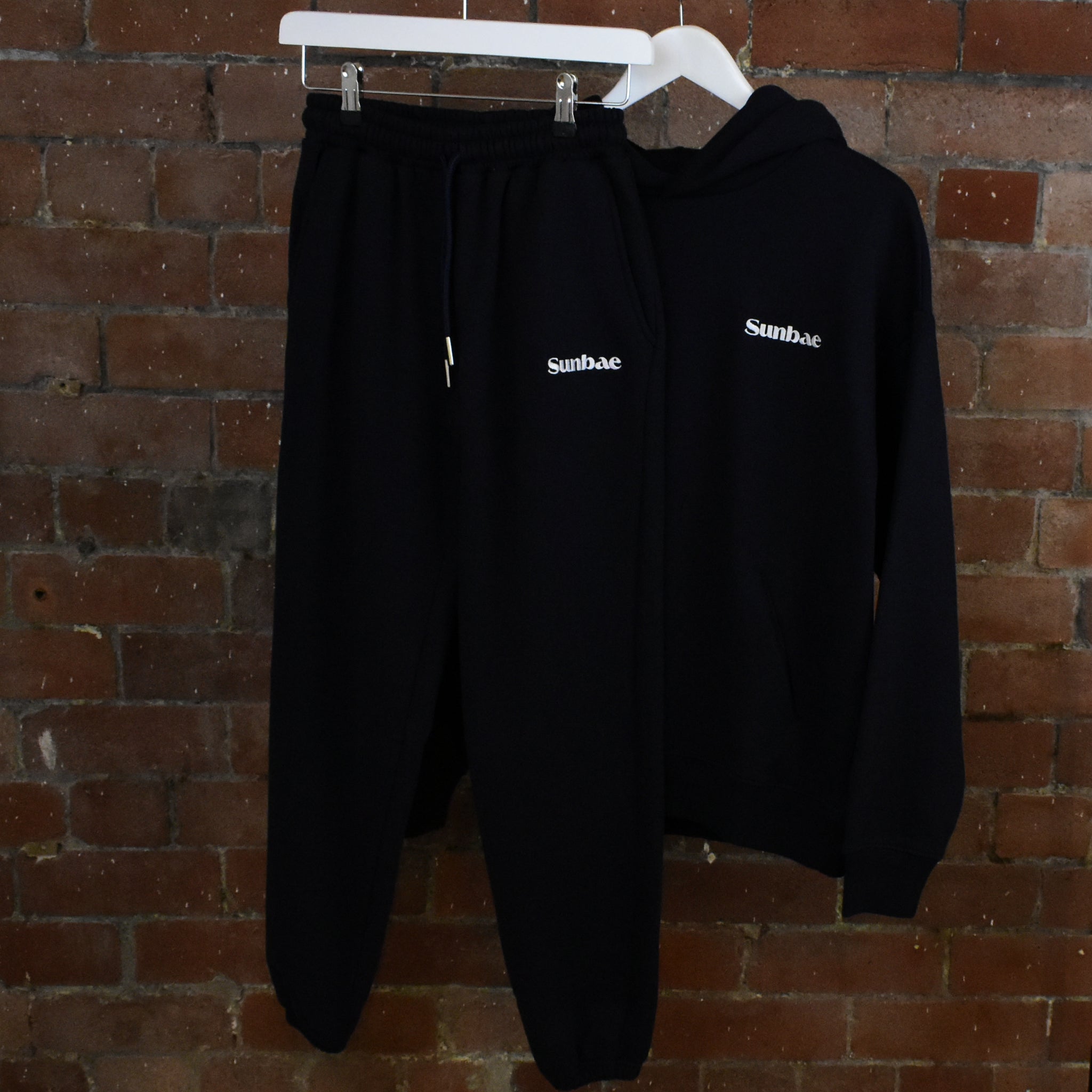 Navy SUNBAE hoodie and Jogger with logo on a hanger against a brick wall 
