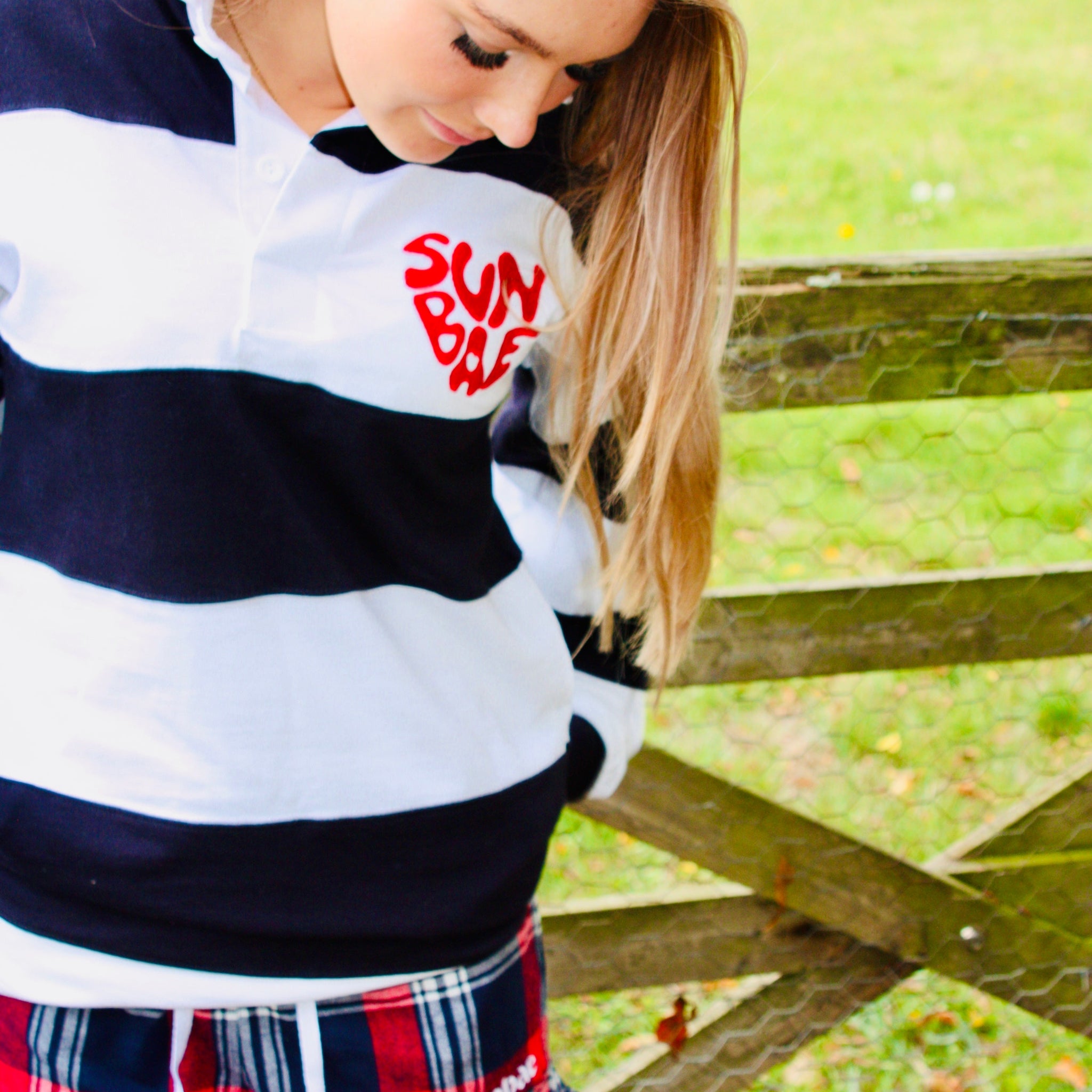 Woman wearing a striped sweater with 'Sun Bum' logo and plaid shorts in an outdoor setting.