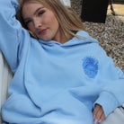 Woman wearing a SUNBAE Tidal Glow light blue hoodie, lying on a hammock.