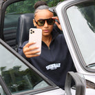 Person taking a selfie in a car wearing sunglasses and a dark hoodie with 'SUNBAE' text.