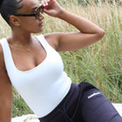 Woman sitting outdoors wearing a white tank top and black leggings with a visible SUNBAE logo.