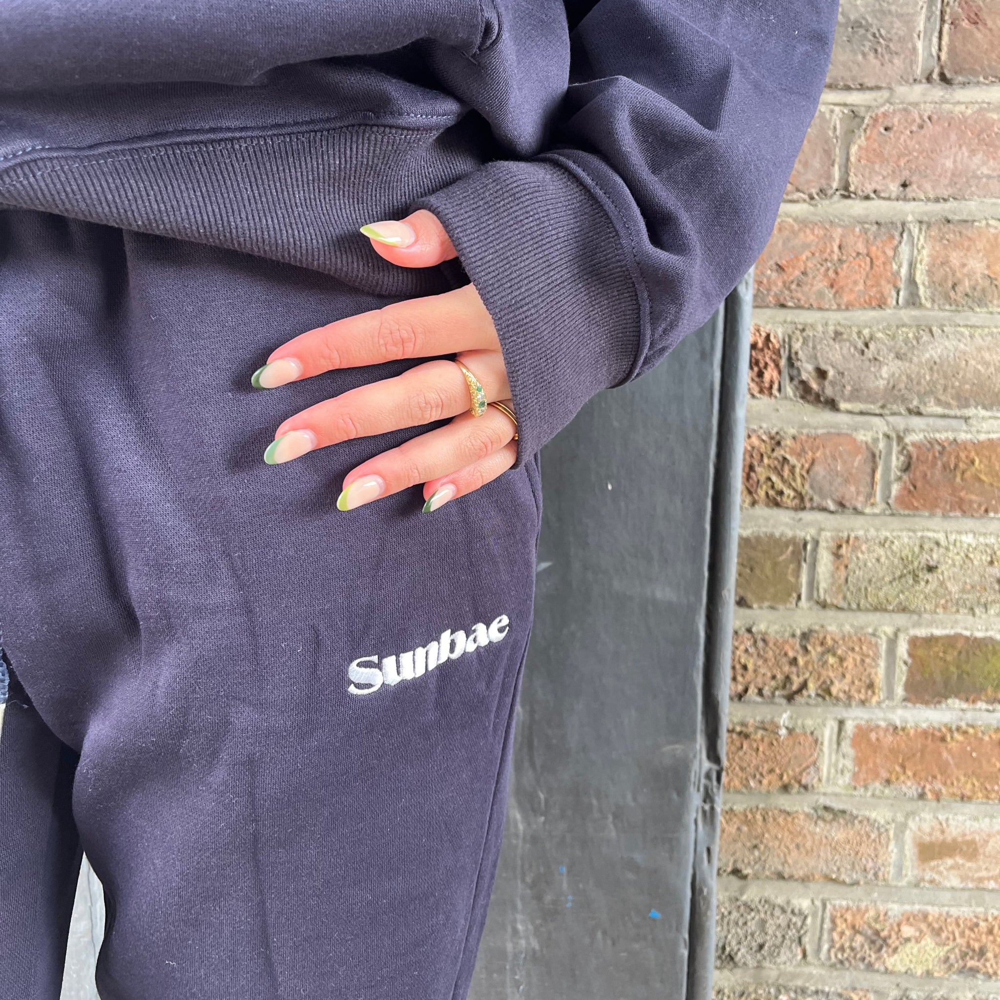 Person wearing a navy blue SUNBAE hoodie and Jogger standing against a brick wall.