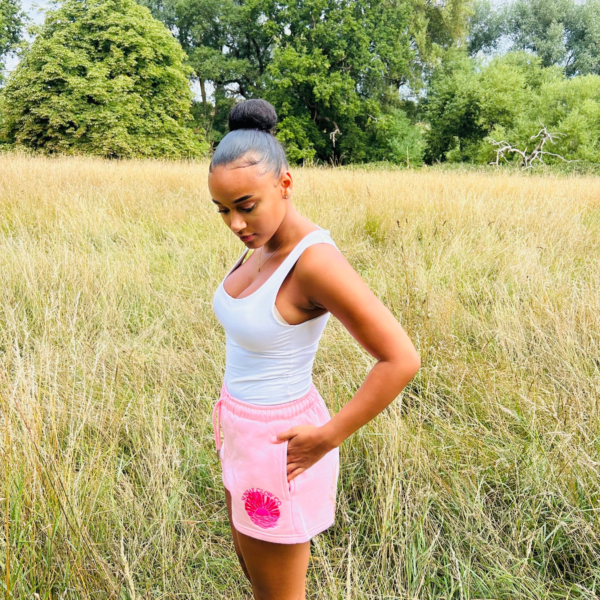 Person wearing a white tank top and pink Sunbae  shorts standing in a grassy field with trees in the background