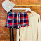 Plaid SUNBAE shorts, beige hoodie, and beanie on a rack against a brick wall.