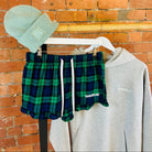 Plaid SUNBAE shorts, beanie, and sweatshirt on a rack against a brick wall.