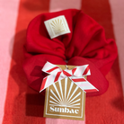 Red SUNBAE Scrunchie with a white tag featuring a design and a gold 'SUNBAE' label on a red and pink striped background.