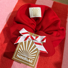 Red SUNBAE scrunchie with a white bow and SUNBAE tag on a red and pink checkered background