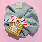 Grey Marl SUNBAE scrunchie with a red and white striped bow SUNBAE tag on a pink surface.