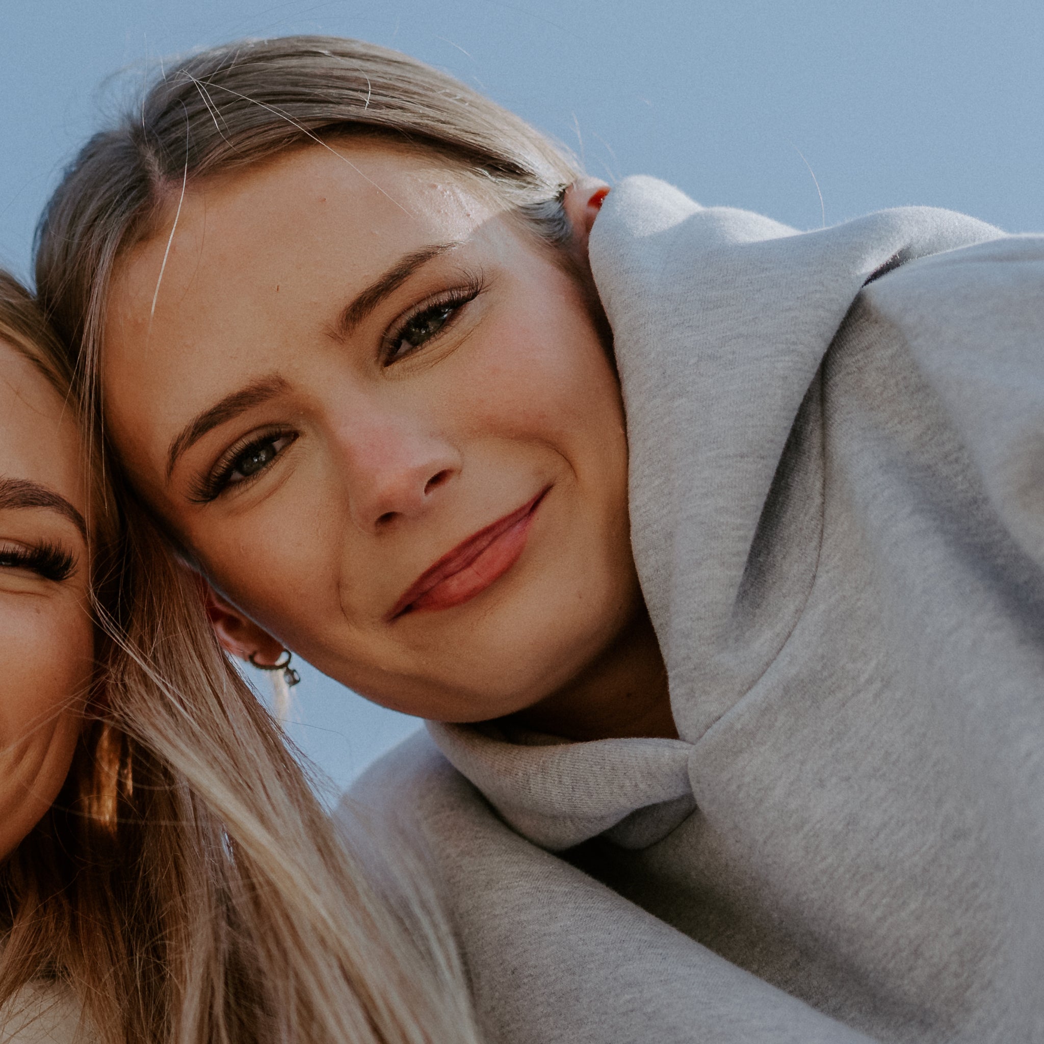 Two women posing closely together with a clear blue sky in the background wearing a SUNBAE grey hoodie