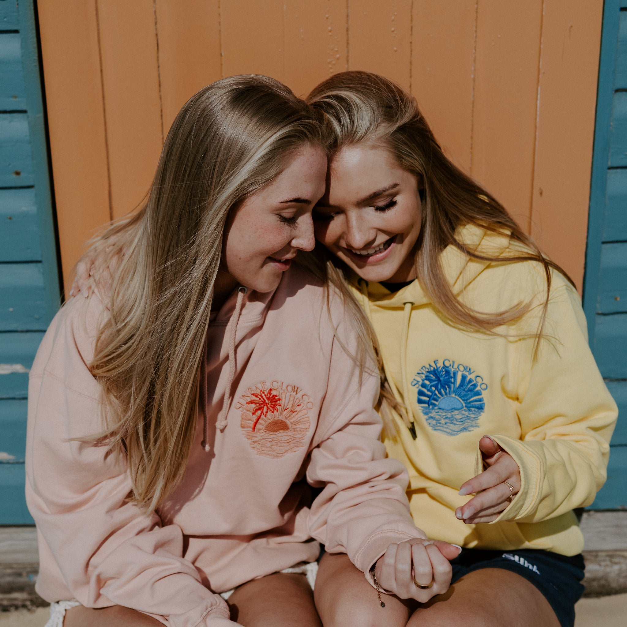 Two women sitting together wearing Sunbae hoodies with visible branding against a colorful wooden background.