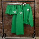 Green SUNBAE hoodie, sweatpants, and shorts set with logo on a rack against a brick wall
