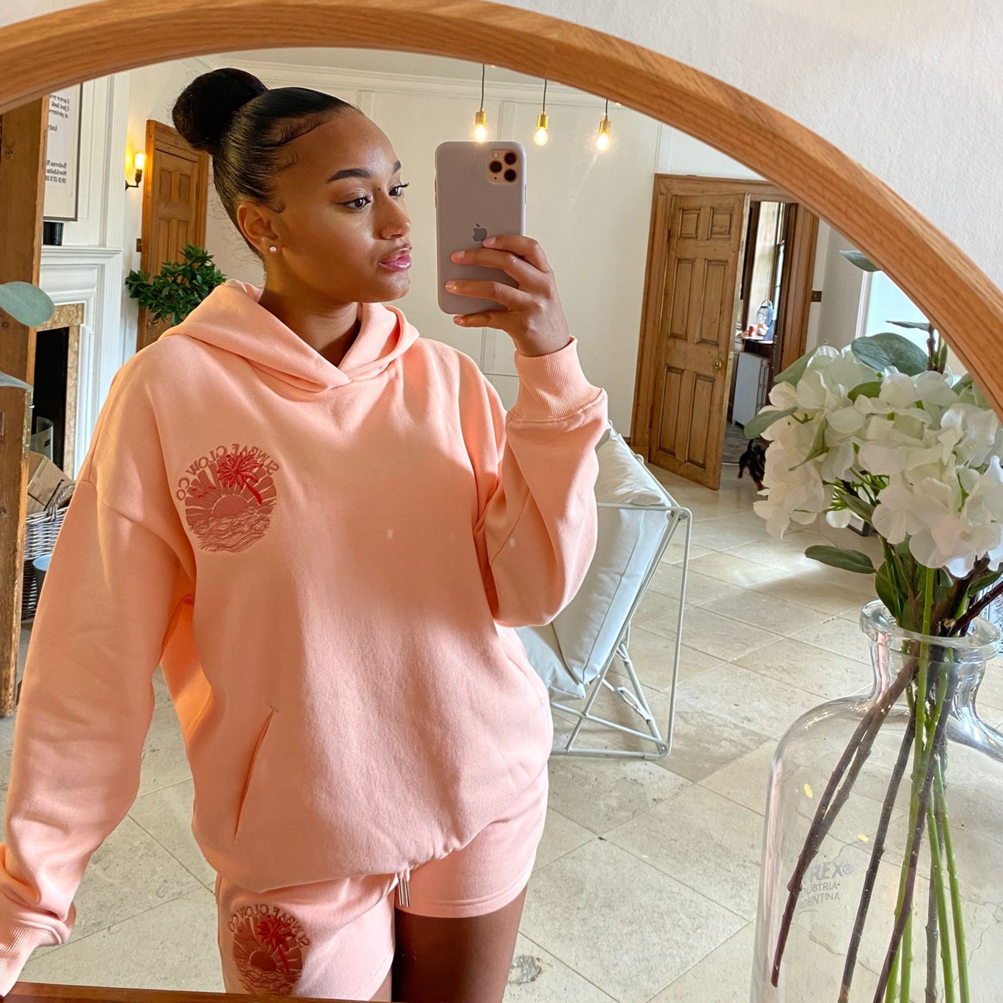 Woman taking a mirror selfie wearing a coral Sunbae hoodie in a home setting.
